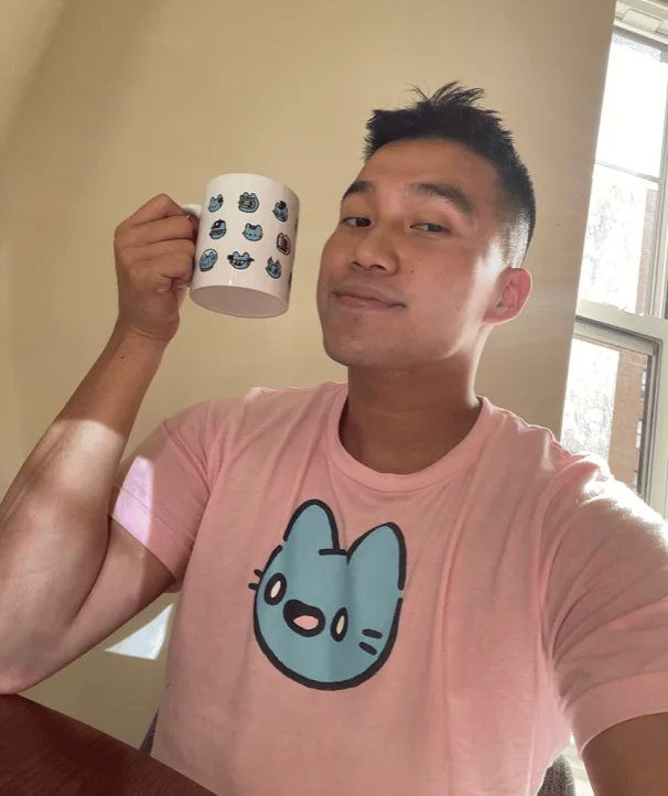 Andrew Wang ( @Andr3w ) smiling holding a Cool Cats mug wearing a pink Cool Cats shirt